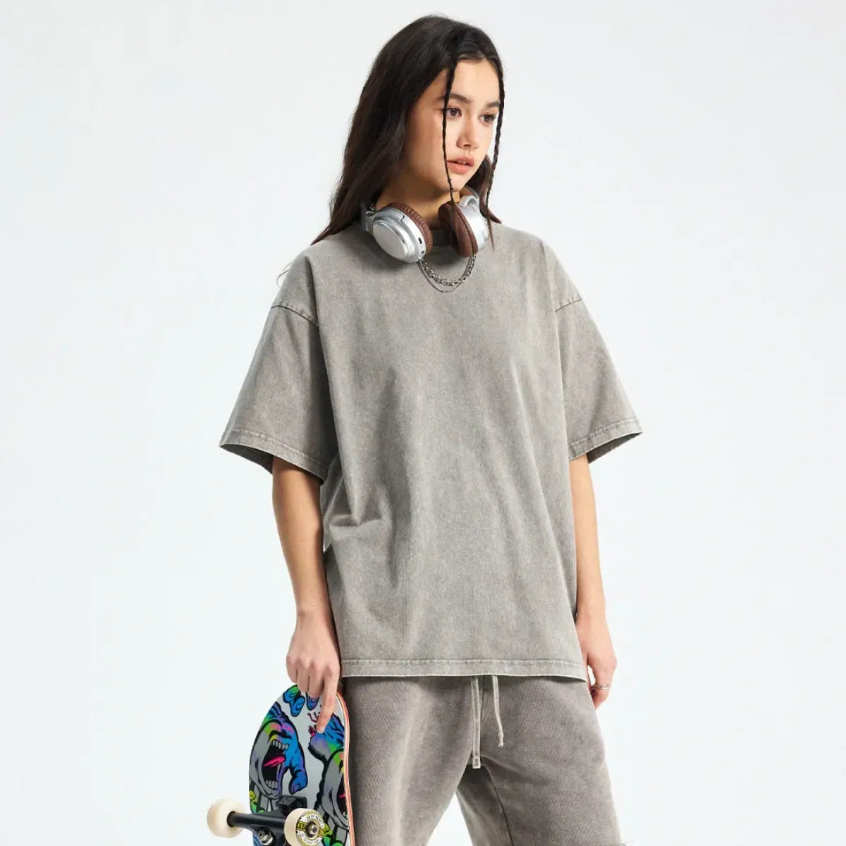 Young person wearing a loose grey t-shirt and sweatpants, headphones around neck, holding a skateboard with a colorful graphic, standing against a plain background.