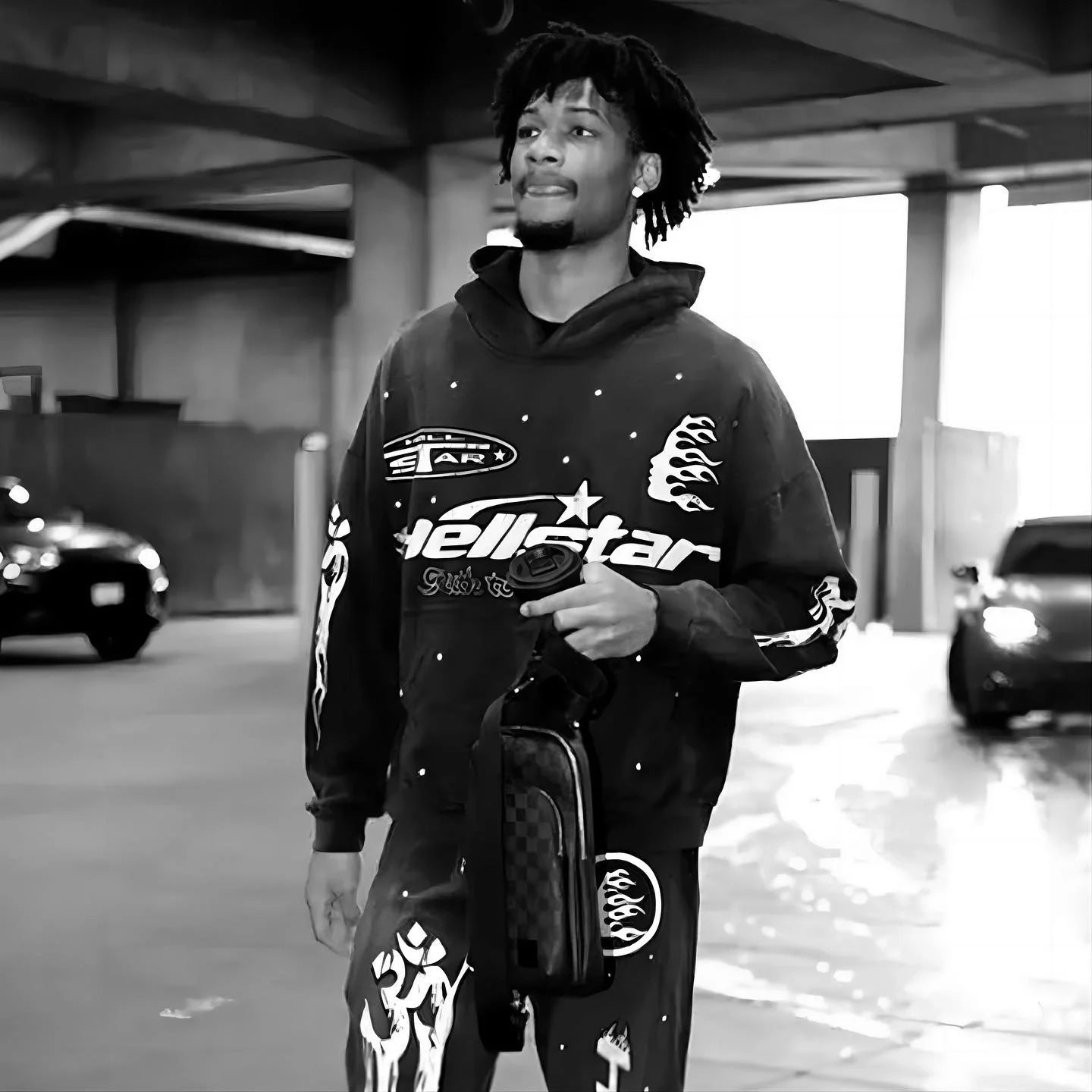 A man wearing a matching graphic hoodie and pants walks in a parking garage, holding a small checkered bag. The image is in black and white.
