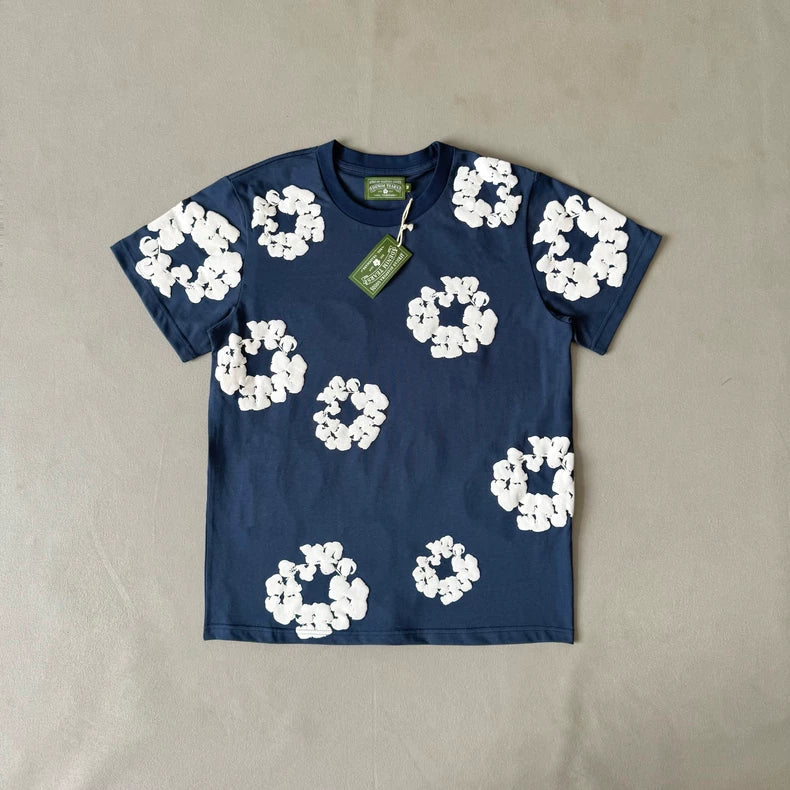 The Denim Tears Navy Cotton Wreath All-Over Print T-Shirt is oversized with bold, irregular white floral graphics on the front and two green tags at the collar. It is displayed flat on a plain, light gray background.