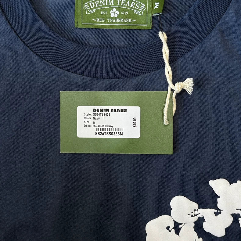 Close-up of the Denim Tears Navy Cotton Wreath All-Over Print T-Shirt, showcasing its white cotton wreath graphic and a green tag detailing style, color, size, and price next to the bold floral design.
