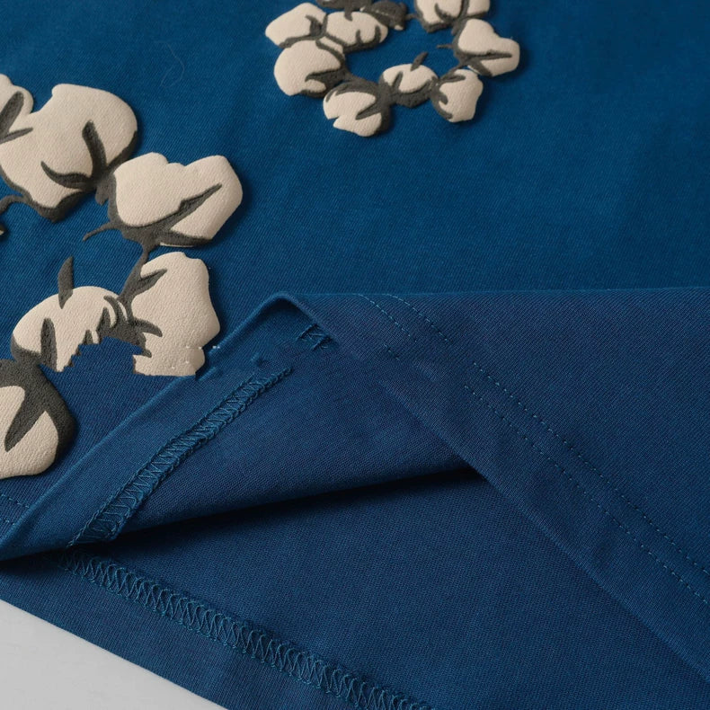A close-up of the Denim Tears Blue Cotton Wreath Allover Print T-Shirt features blue fabric with beige and brown floral patterns, detailed stitching, and a folded corner, echoing its signature cotton wreath graphic.