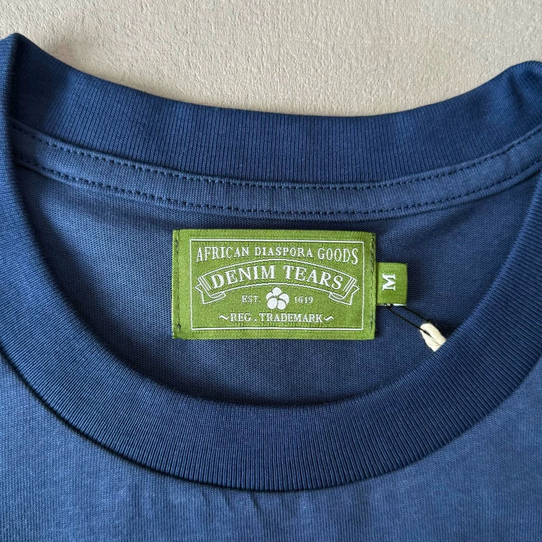 A close-up of the Denim Tears Navy Cotton Wreath All-Over Print T-Shirt shows its blue collar with a green “African Diaspora Goods DENIM TEARS” label and an attached “M” size tag, highlighting the detail of this oversized tee.
