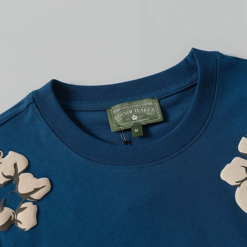 A close-up of the Denim Tears Blue Cotton Wreath Allover Print T-Shirt featuring short sleeves, a green label, and beige floral appliqué designs near the shoulders.