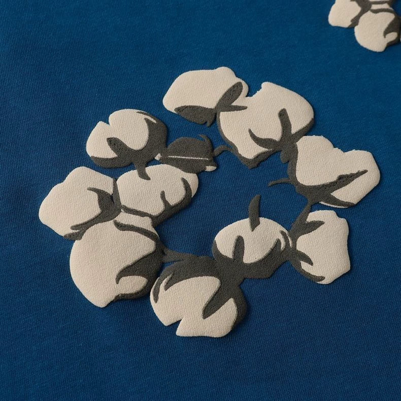 The Denim Tears Blue Cotton Wreath Allover Print T-Shirt features stylized beige cotton bolls with dark gray stems, arranged in a circle on a blue background, inspired by signature wreath motifs.
