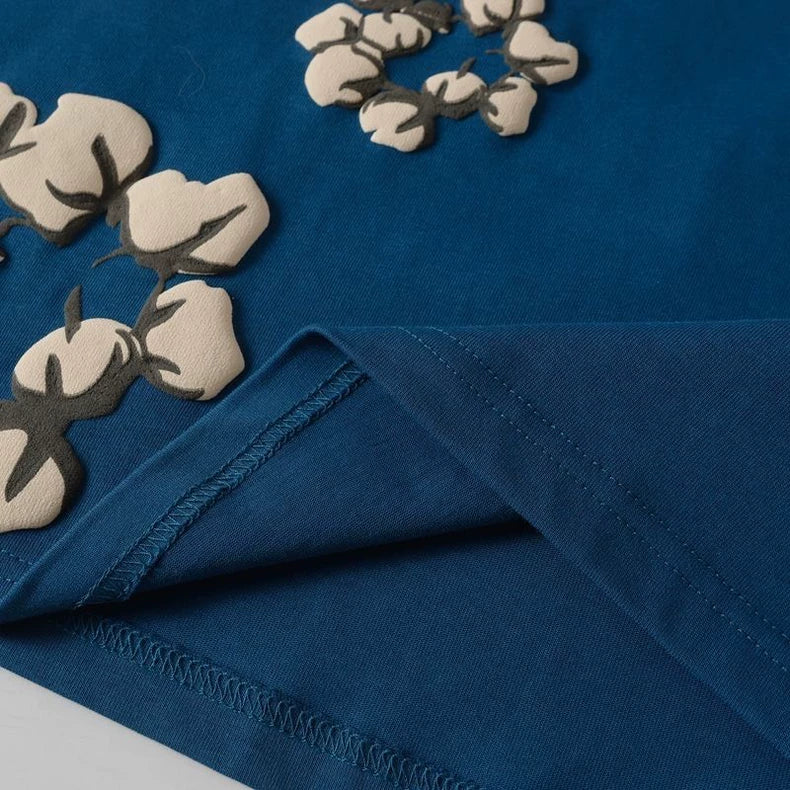 A close-up of the Denim Tears Blue Cotton Wreath Allover Print T-Shirt shows blue fabric with beige and dark brown floral applique details. A fold in the fabric reveals edge stitching.