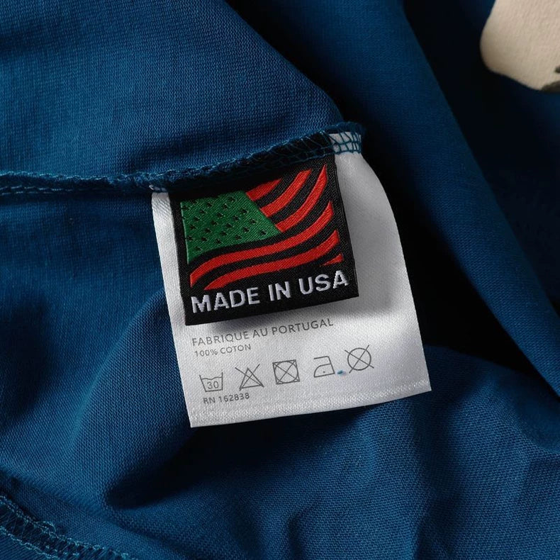A tag on the Denim Tears Blue Cotton Wreath Allover Print T-Shirt sewn onto blue fabric has a stylized American flag, "MADE IN USA," "FABRIQUé AU PORTUGAL," "100% COTON," care symbols, and an RN number.
