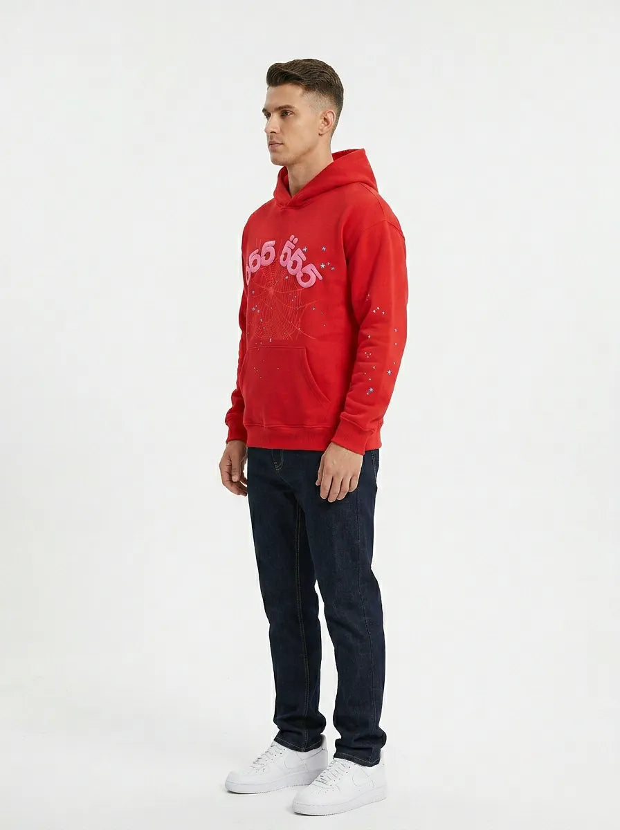 A man stands against a plain background in a Red 555 Sp5der Hoodie – 440GSM Heavyweight Sp5der 555 Web Oversized Pullover, styled with streetwear-inspired prints, dark jeans, and white sneakers.