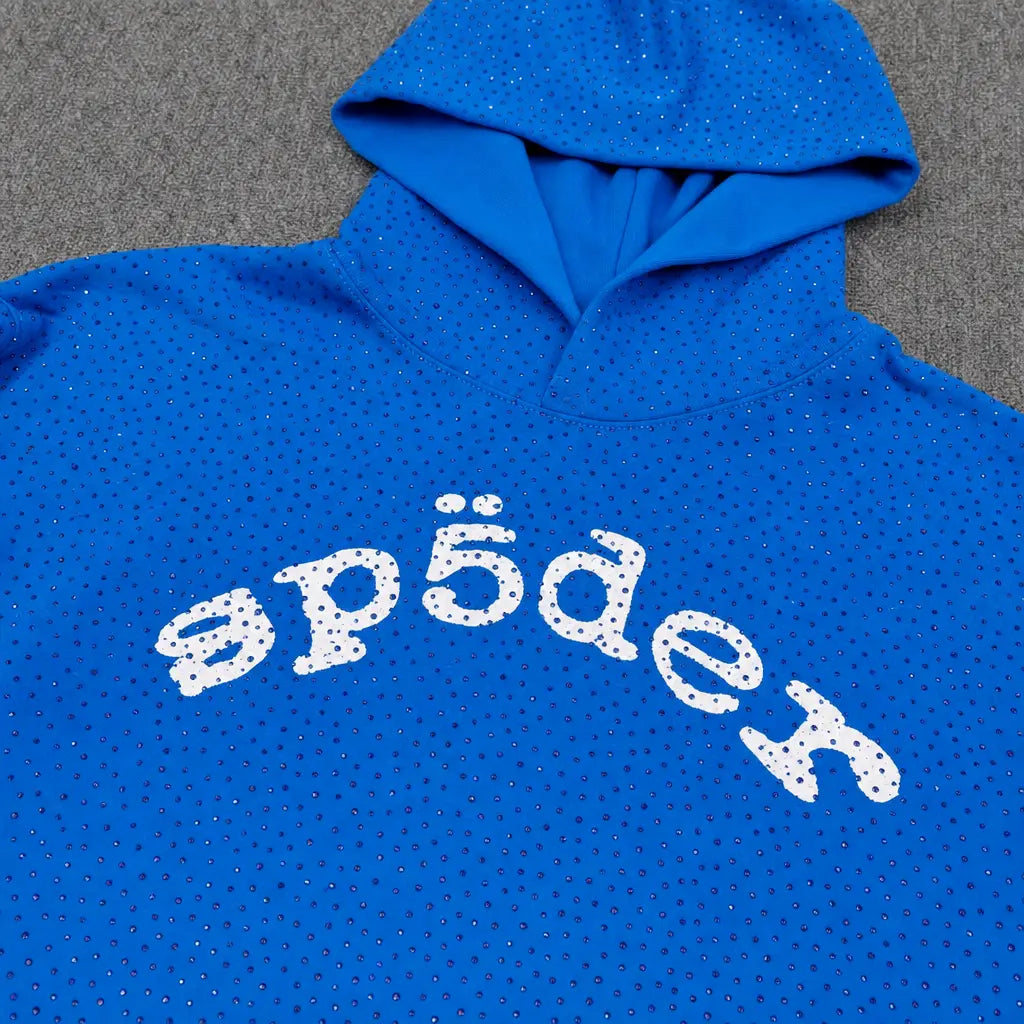 The Sp5der Blue Rhinestone All-Over Arch Logo Hoodie features small rhinestones and a white “sp5der” arch logo across the chest, creating a standout streetwear look. It’s displayed on a gray surface.