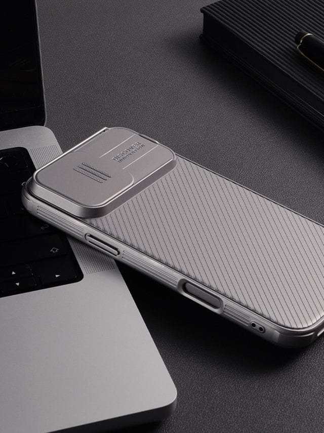 Close-up of a silver iPhone 16 Pro Max Camera Slider Case with anti-scratch, shockproof, and slim fit protection, featuring a textured back, partially placed on a laptop's keyboard. A pen and closed notebook are visible in the background.