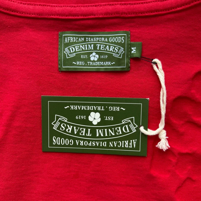 The Denim Tears Red Cotton Wreath Allover Print T-Shirt features a crew neck, green label inside the collar, and a matching green tag attached by white string. Both labels read: African Diaspora Goods, Est. 1619, Reg. Trademark.