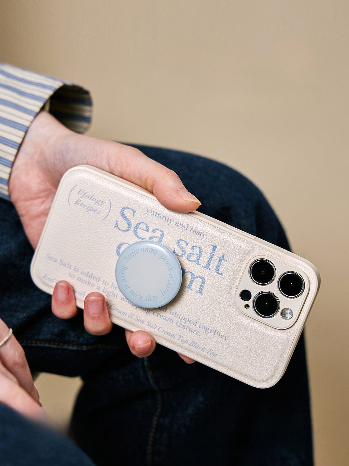 A person is holding an iPhone 16 Pro Max with a "Sea Salt Cream" MagSafe case, featuring a minimalist textured design, and a blue phone grip.