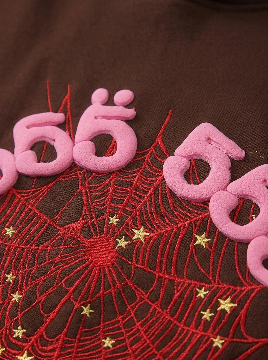 Close-up of the 555 Brown Spider Hoodie – Sp5der 555 Web Brown/Pink, a heavyweight oversized pullover featuring pink raised 555 text and a red embroidered spider web with yellow stars on soft brown fabric.