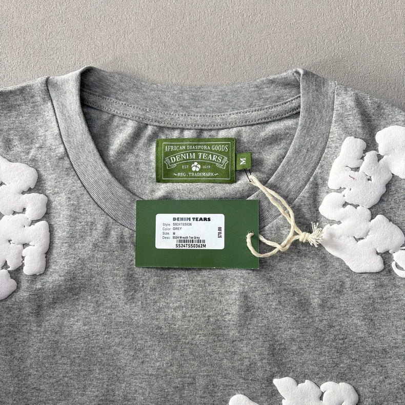 The Denim Tears Grey Cotton Wreath Allover Print T-Shirt - 2 features a heather grey cotton base with white wreath graphics and floral appliqués, plus a green “Denim Tears” collar label and matching green and white product tag.