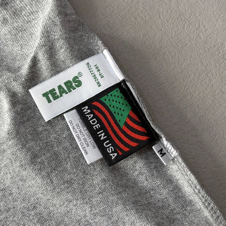 Close-up of a heather grey Denim Tears Grey Cotton Wreath Allover Print T-Shirt - 2 collar showing three tags: TEARS?, MADE IN USA with a stylized flag, and an M tag for medium size.