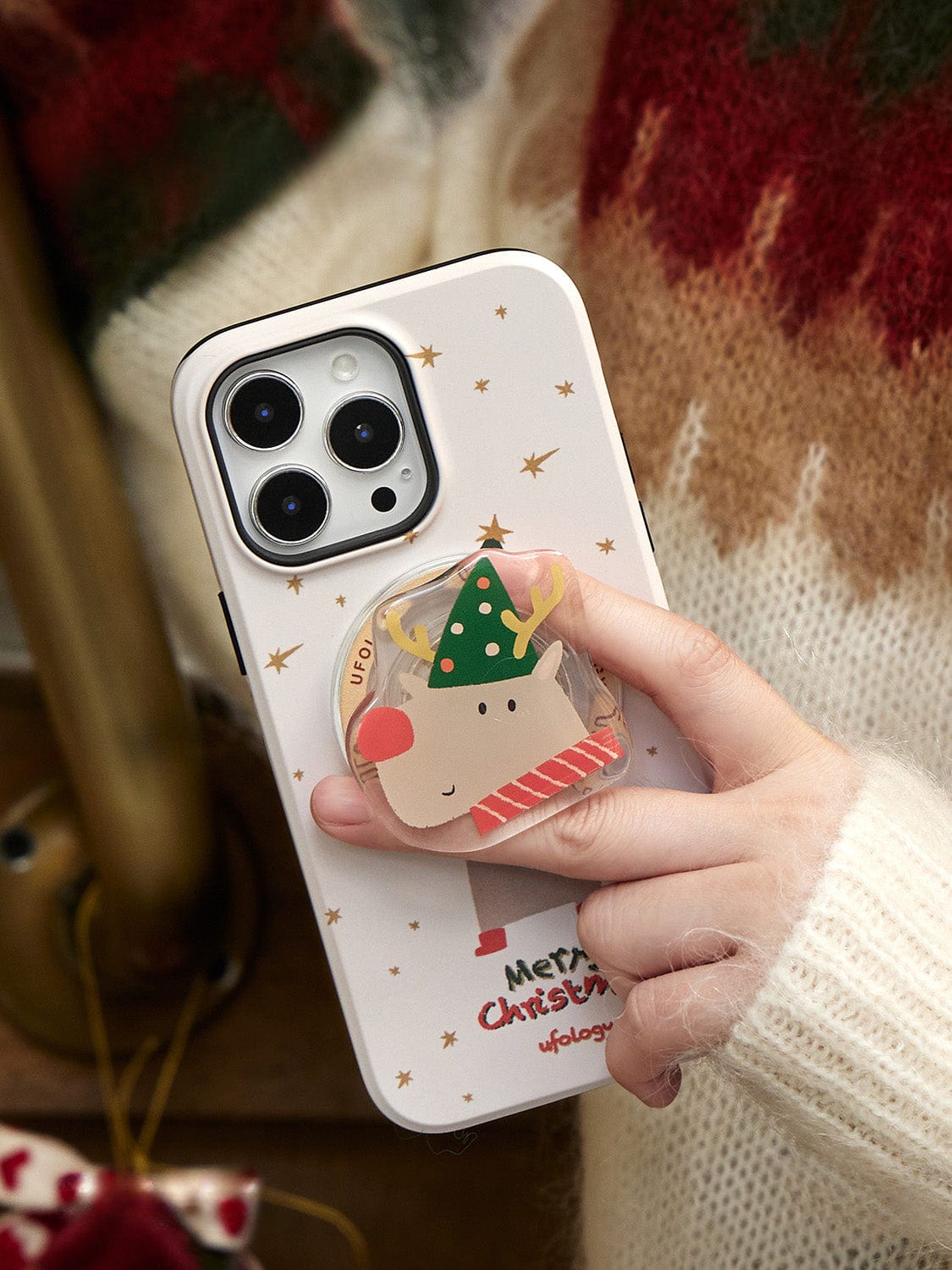 A person holding a smartphone with a Rudolph Joy MagSafe iPhone 16 Pro Max Case, featuring antlers, a green hat, and "Merry Christmas" text.