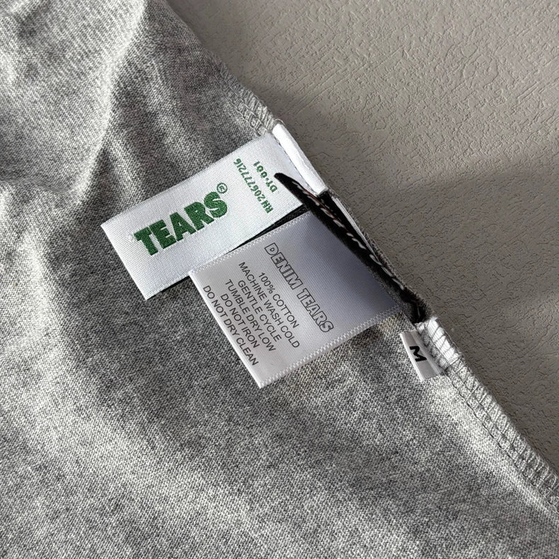 Close-up of tags on a Denim Tears Grey Cotton Wreath Allover Print T-Shirt - 2 in heather grey. Tags feature TEARS? in green and DENIM TEARS, along with sizing, material, and care info printed in black.
