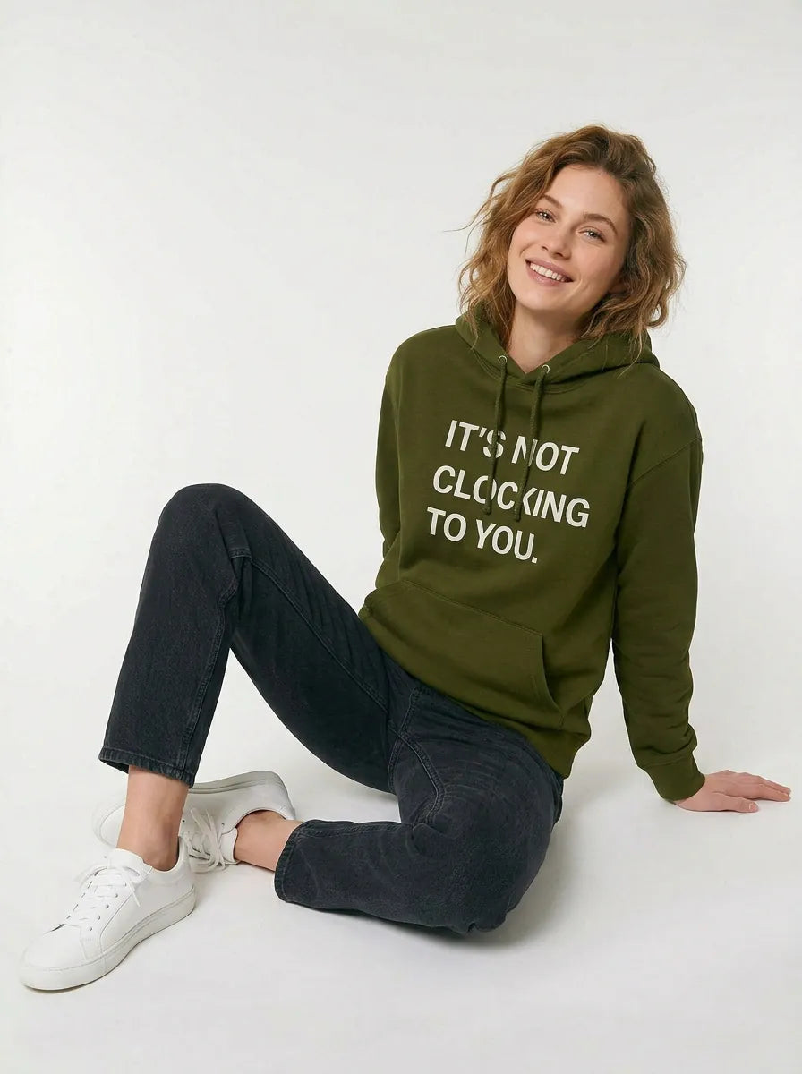 A person sits on the floor wearing a green "Its Not Clocking To You Hoodie - 360GSM Heavyweight 100% Cotton Graphic Pullover Hoodie" and black jeans, smiling at the camera.