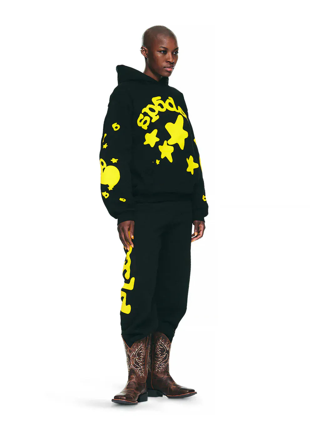 A person wears the Black and Yellow Spider Hoodie with Star Lettering Print – Comfort Fit Pullover Hoodie for Everyday Wear, paired with pants featuring bold yellow graphics and brown cowboy boots, standing against a plain white background.