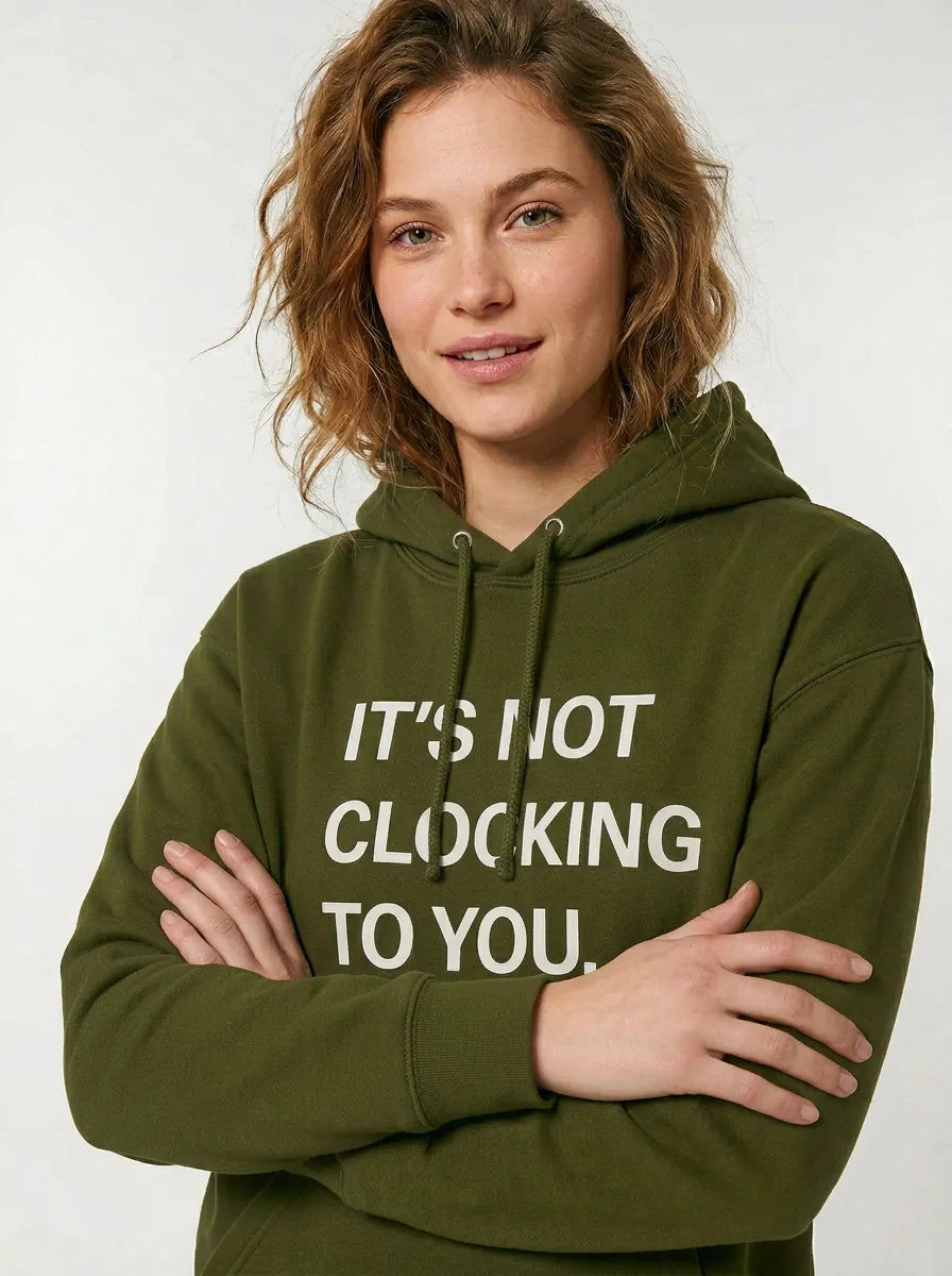 A person with wavy light brown hair stands with arms crossed, wearing the Its Not Clocking To You Hoodie—a 360GSM heavyweight 100% cotton graphic pullover—while looking at the camera.
