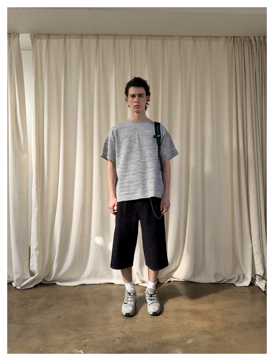 Model wearing a gray striped short-sleeve T-shirt with dark shorts and sneakers, standing in front of a cream curtain