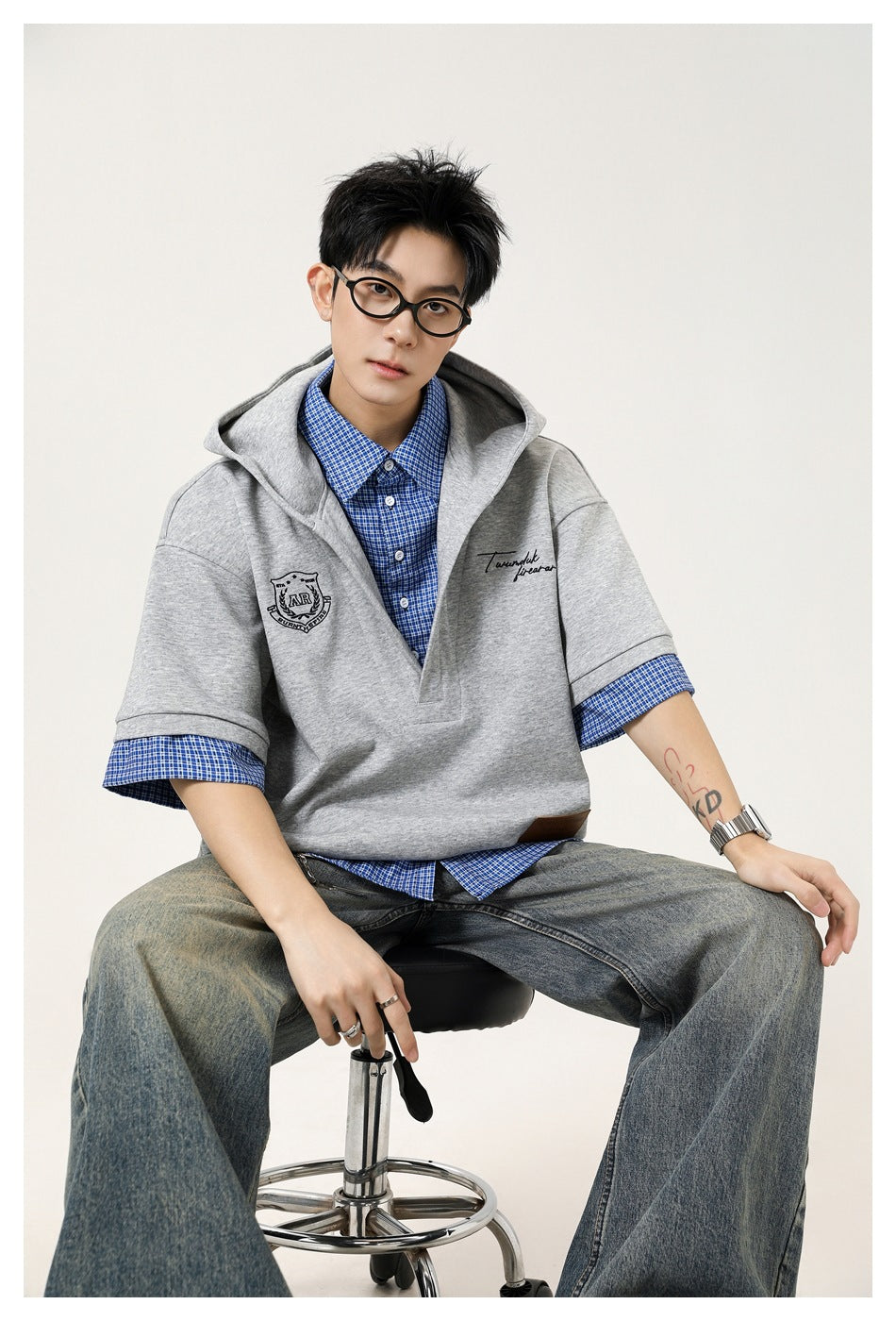 Seated model wearing a light gray hooded pullover over a blue checked shirt, shown from the waist up.