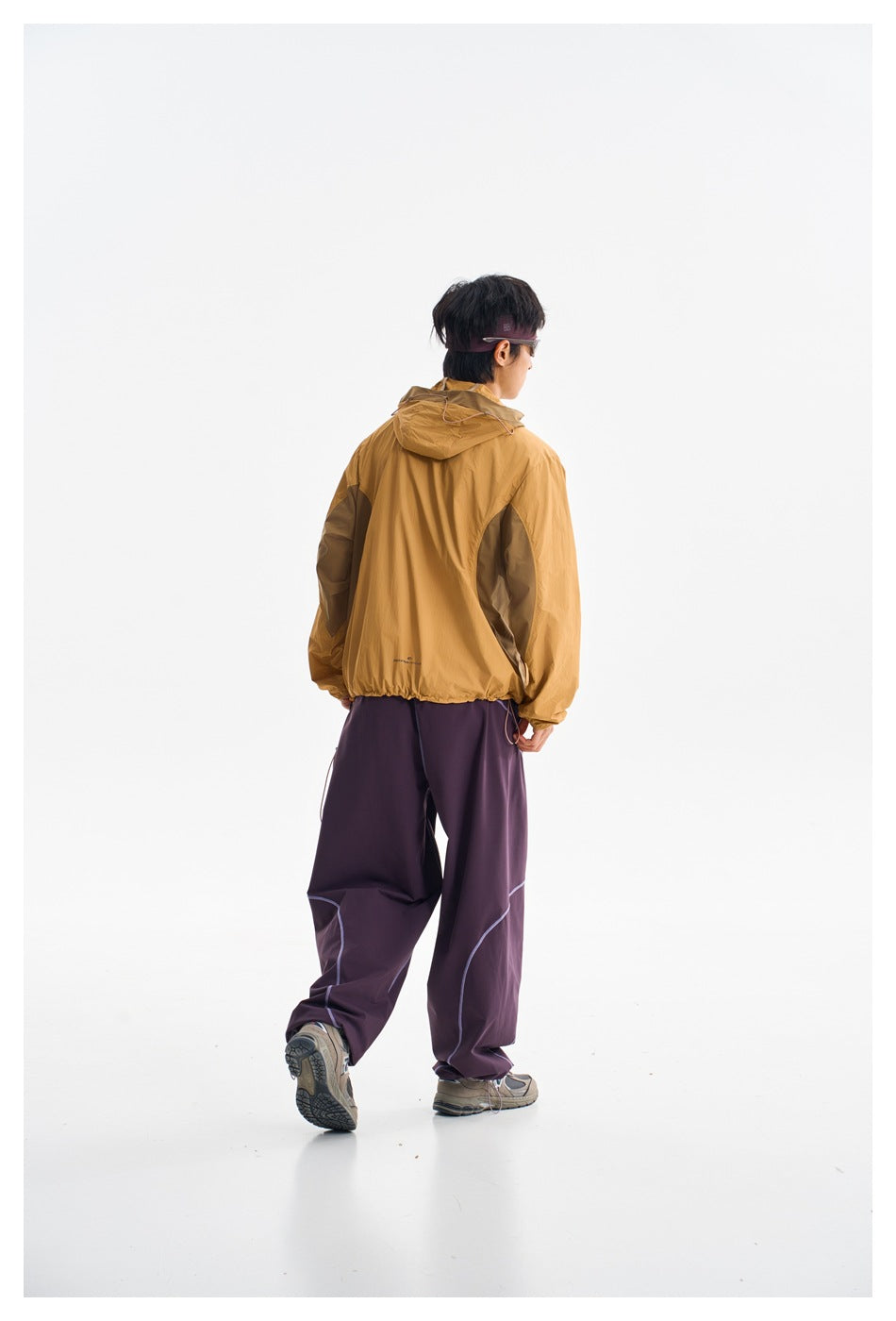 Rear view of a model wearing a mustard hooded jacket and loose plum pants with pale seam detailing.