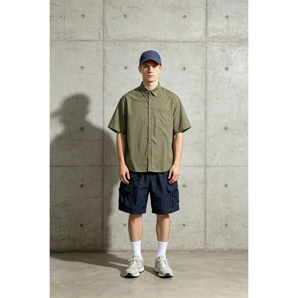 Model wearing an olive short-sleeve button-up shirt with a chest pocket, navy cargo shorts, and a navy cap.