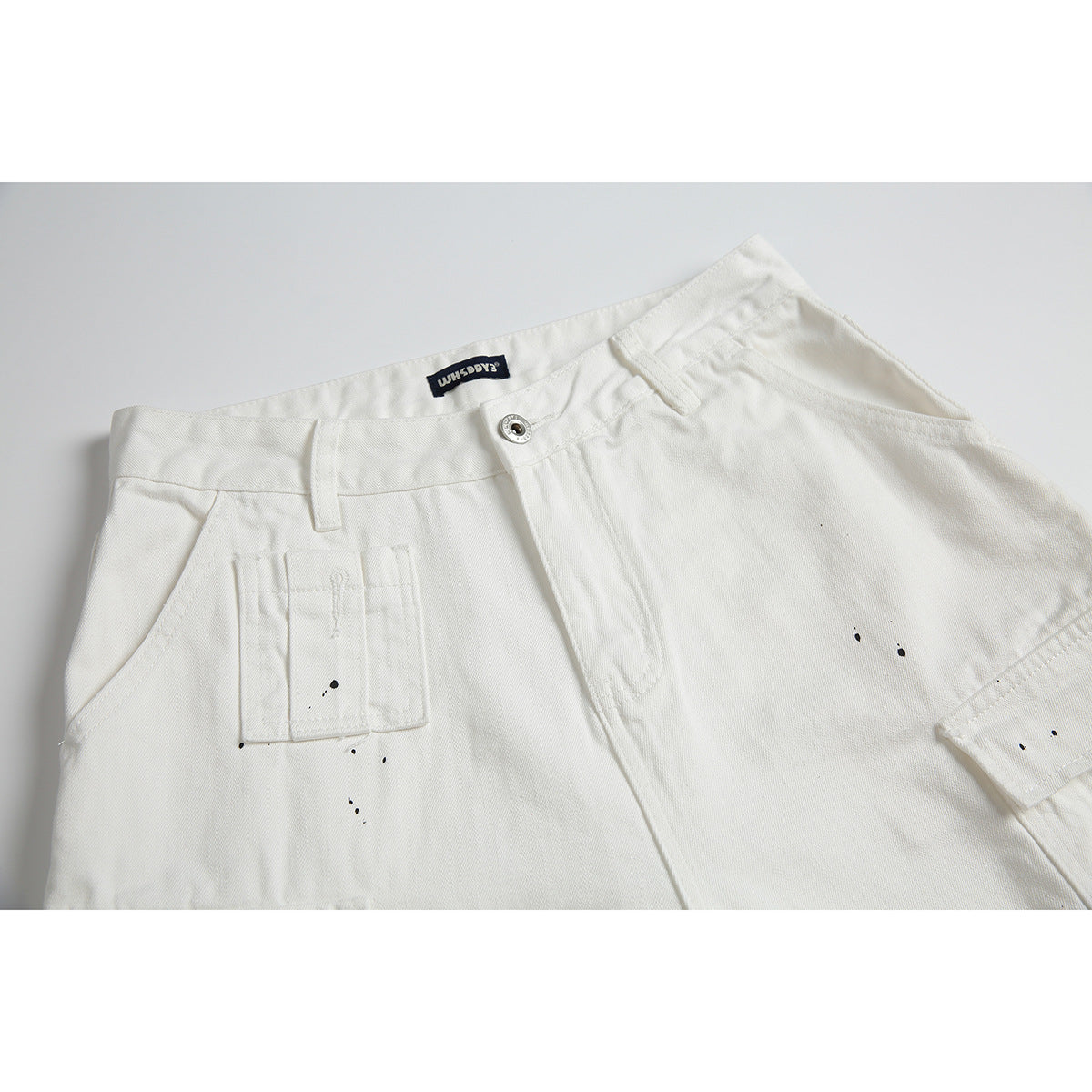 Close-up of white pants front with a button waist, small utility pocket, and scattered black specks.