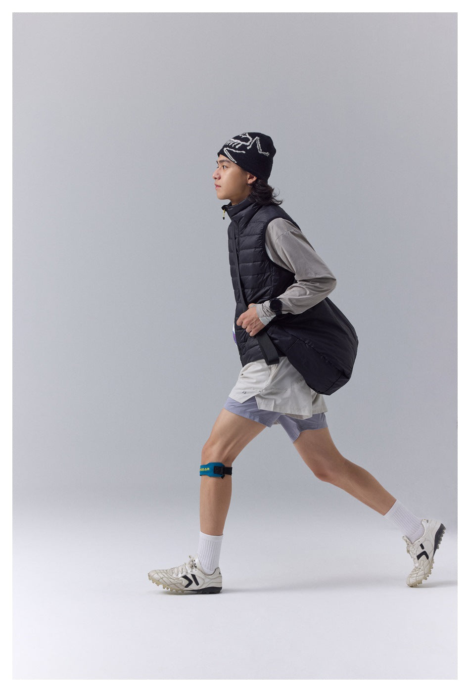 Side view of a model moving in a black quilted vest with a large black bag, light gray top, and athletic shorts.