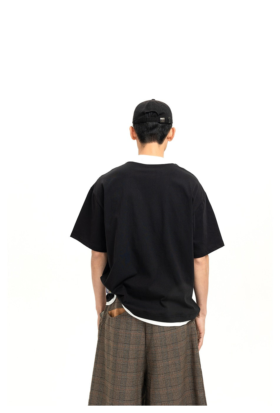 Rear view of a model wearing a loose black short-sleeve top over a white shirt, with plaid trousers.