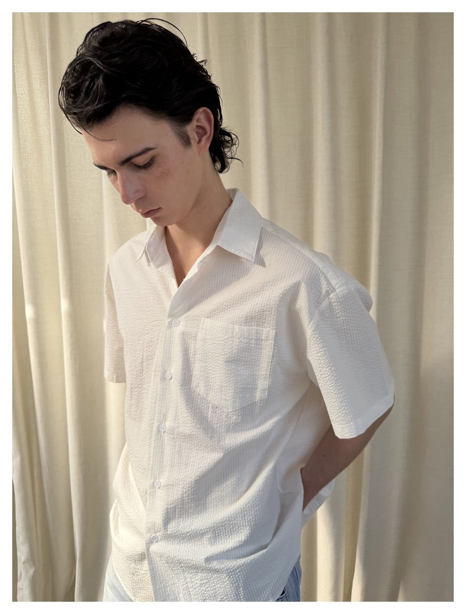 Close-up of a model in a white crinkled short-sleeve shirt with chest pocket, looking down, cream curtain background