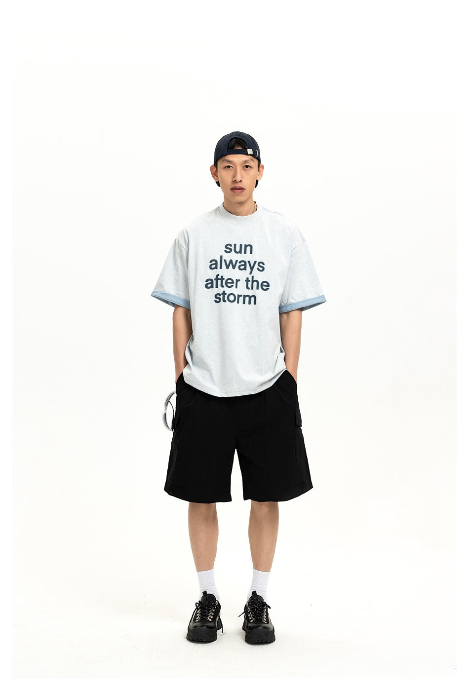 Full-length front view of a model in a light gray slogan T-shirt with blue sleeve cuffs and black shorts.