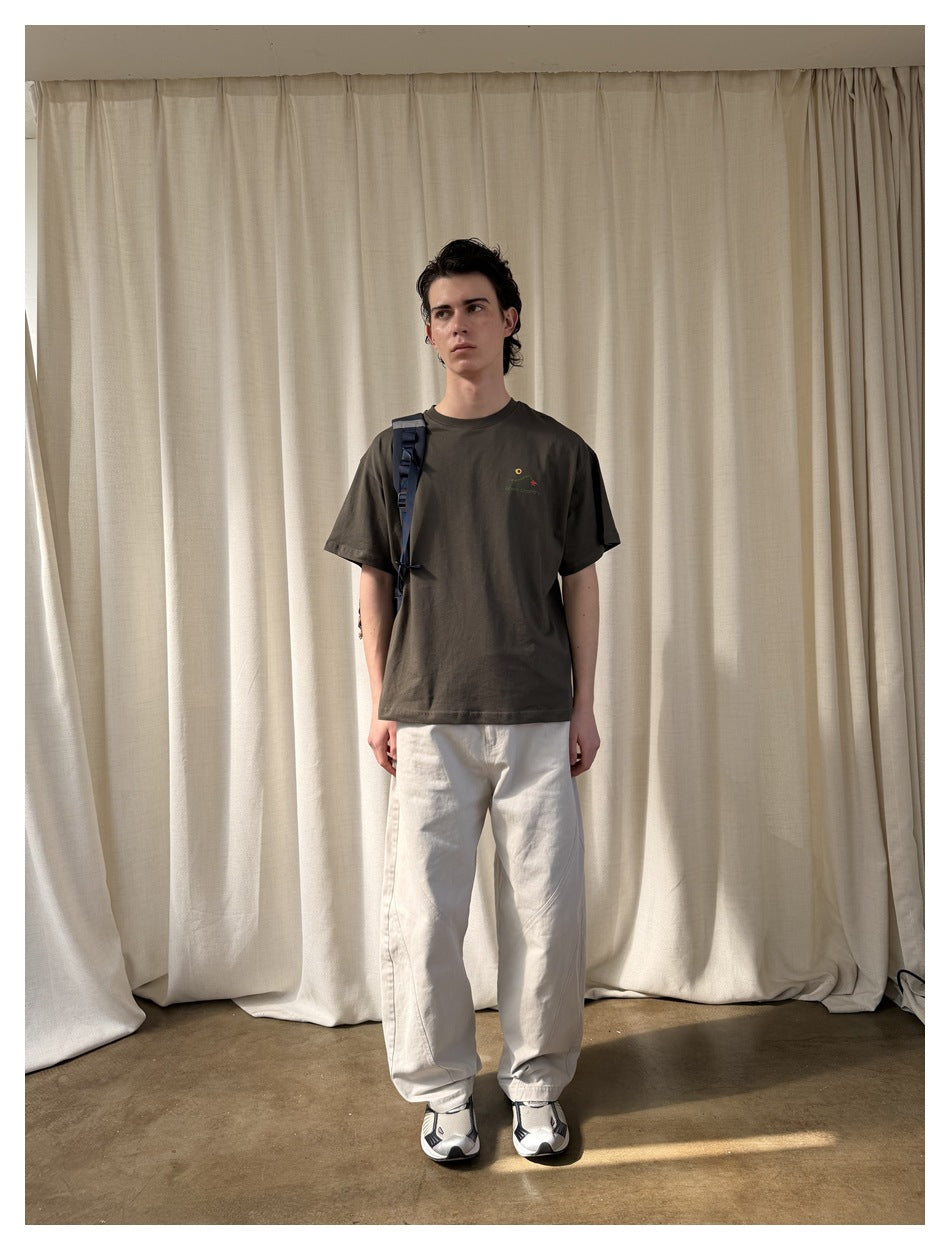 Model wearing a dark olive T-shirt with a small chest graphic and loose white pants.