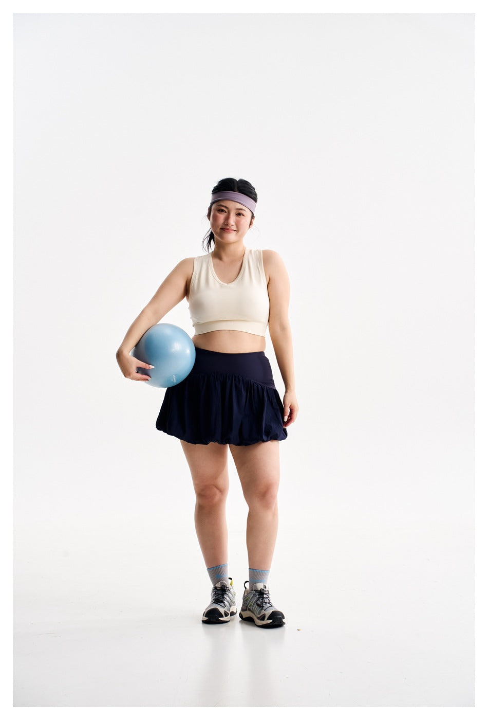 Woman wearing an ivory crop top and navy bubble-hem athletic skirt, holding a blue exercise ball at her side.