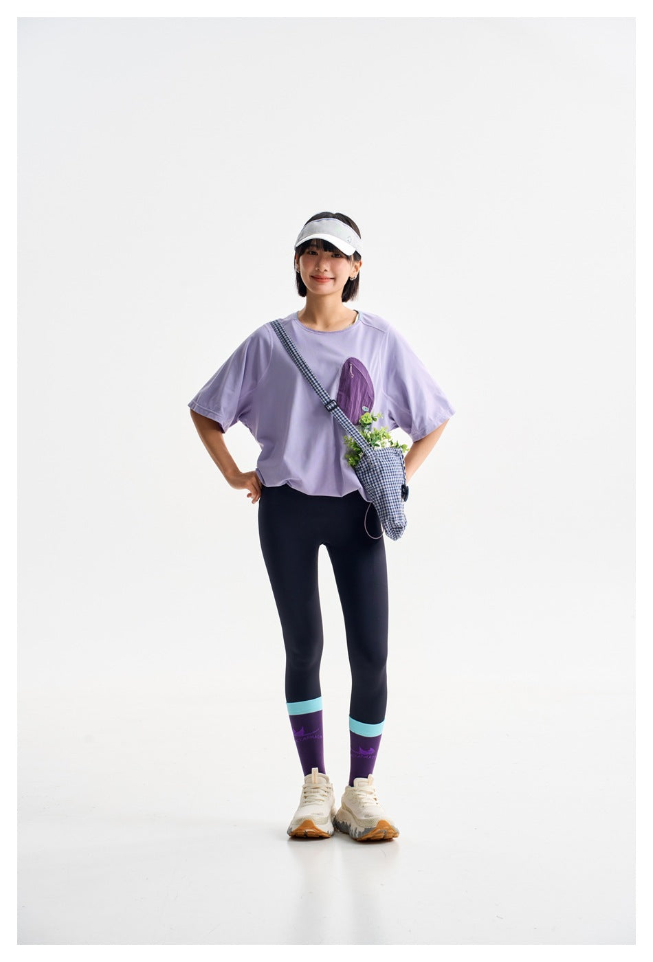 Model posing in a lavender short-sleeve top and dark leggings with purple socks and a gingham crossbody bag.
