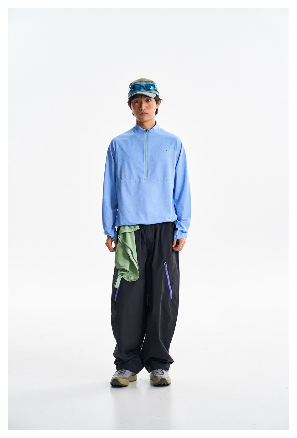 Model wearing a light blue quarter-zip pullover with black pants and a green garment draped at the waist, front view.