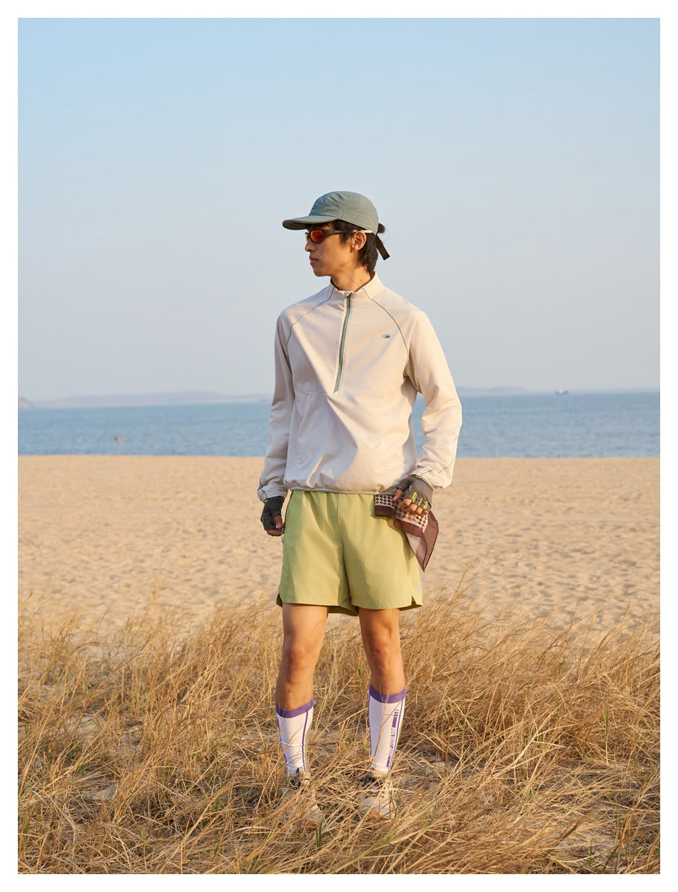 Man standing on a beach in a cream quarter-zip top, olive shorts, cap, sunglasses and tall socks.