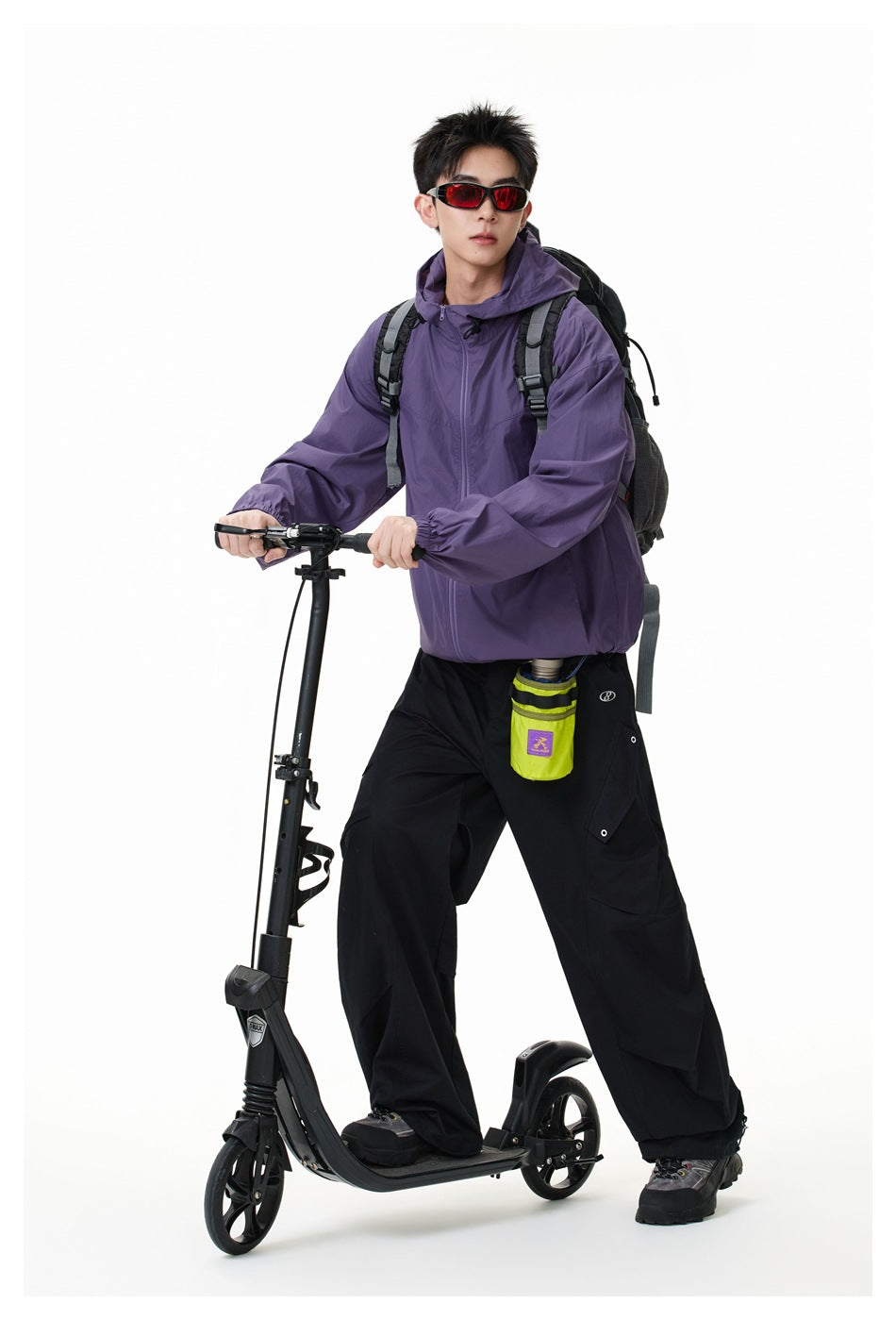Model wearing a purple hooded zip jacket and loose black trousers while standing over a scooter with a backpack.