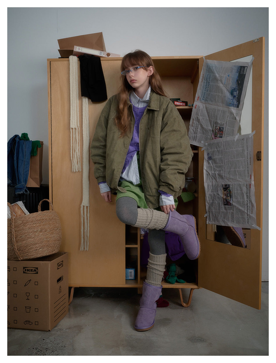 Model wearing an army green zip-front jacket over a striped shirt and purple cardigan, posed leaning against a wooden