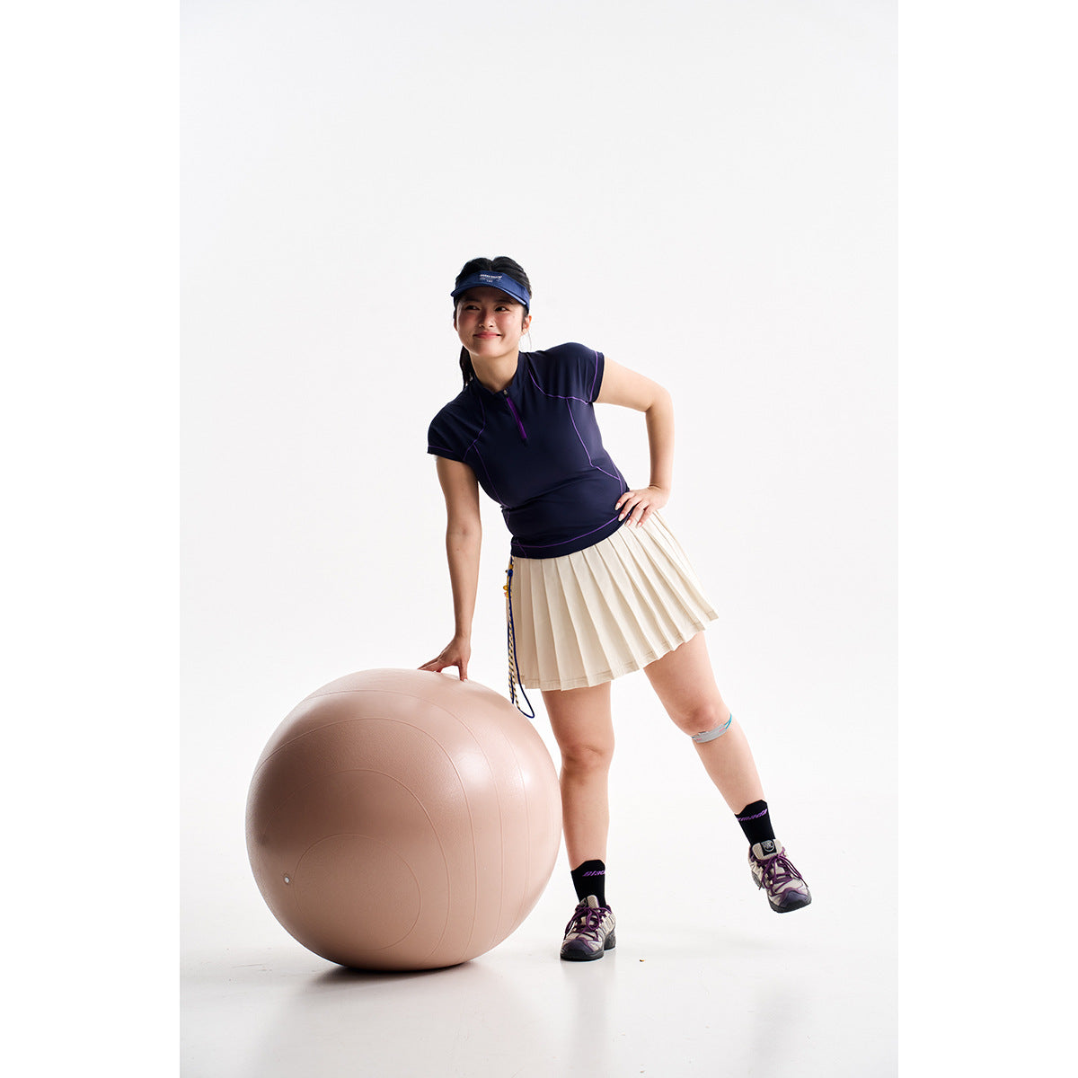 Model wearing a navy zip-neck athletic top and ivory pleated skirt beside a large exercise ball.