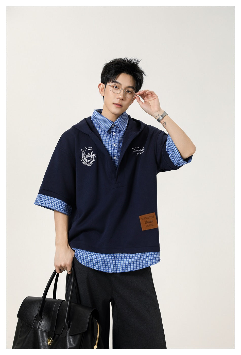 Model in navy layered hooded top with blue check shirt layer, holding a black bag and touching glasses