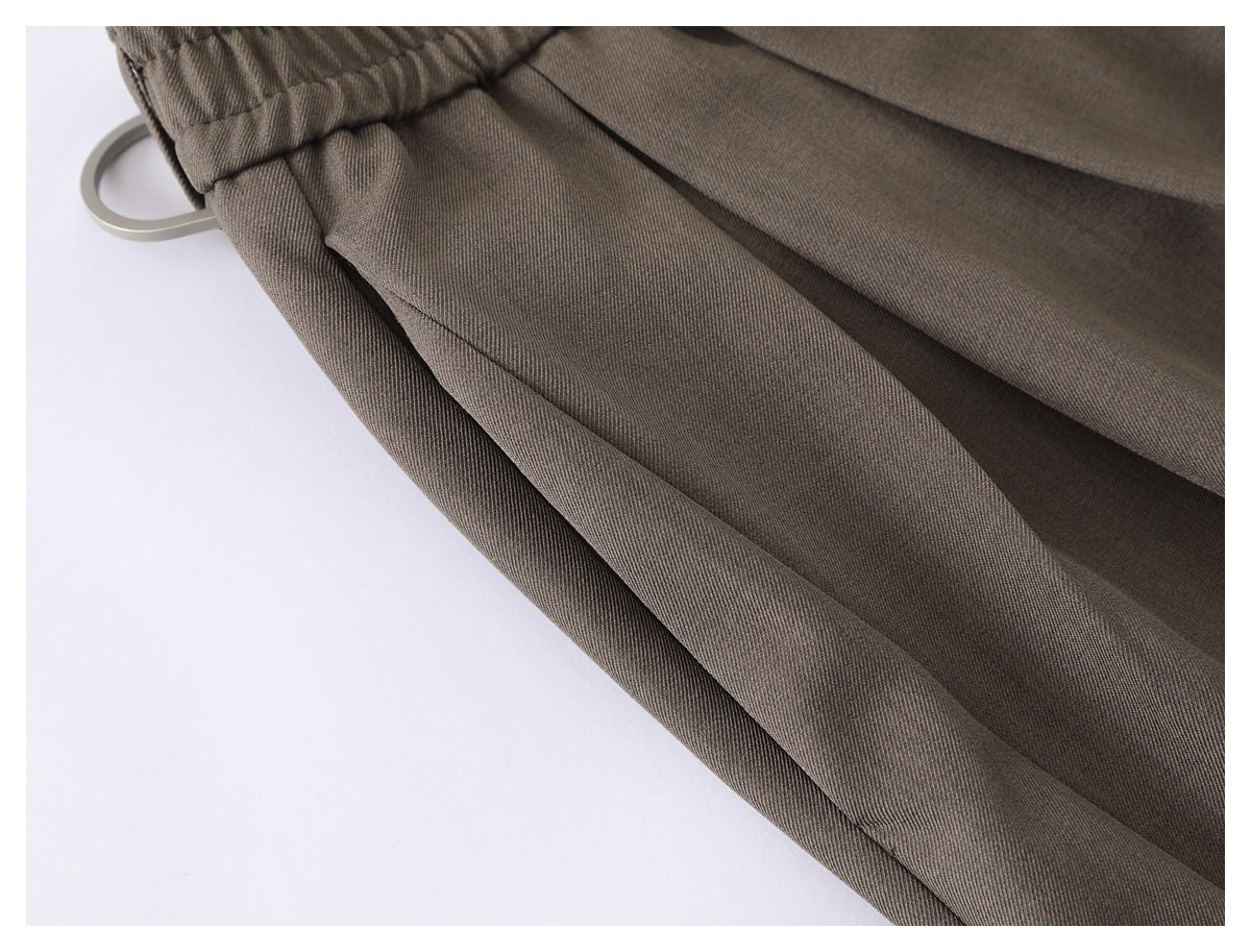 Close-up of taupe trousers showing the gathered elastic waistband and a metal belt ring.