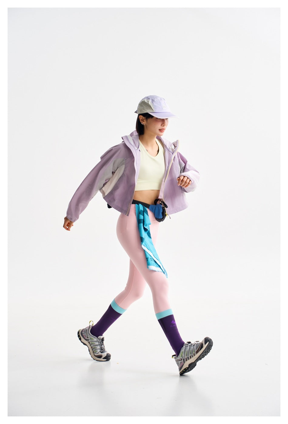 Model walking in a lilac cropped hooded jacket, pale crop top, pink leggings, and lavender cap.