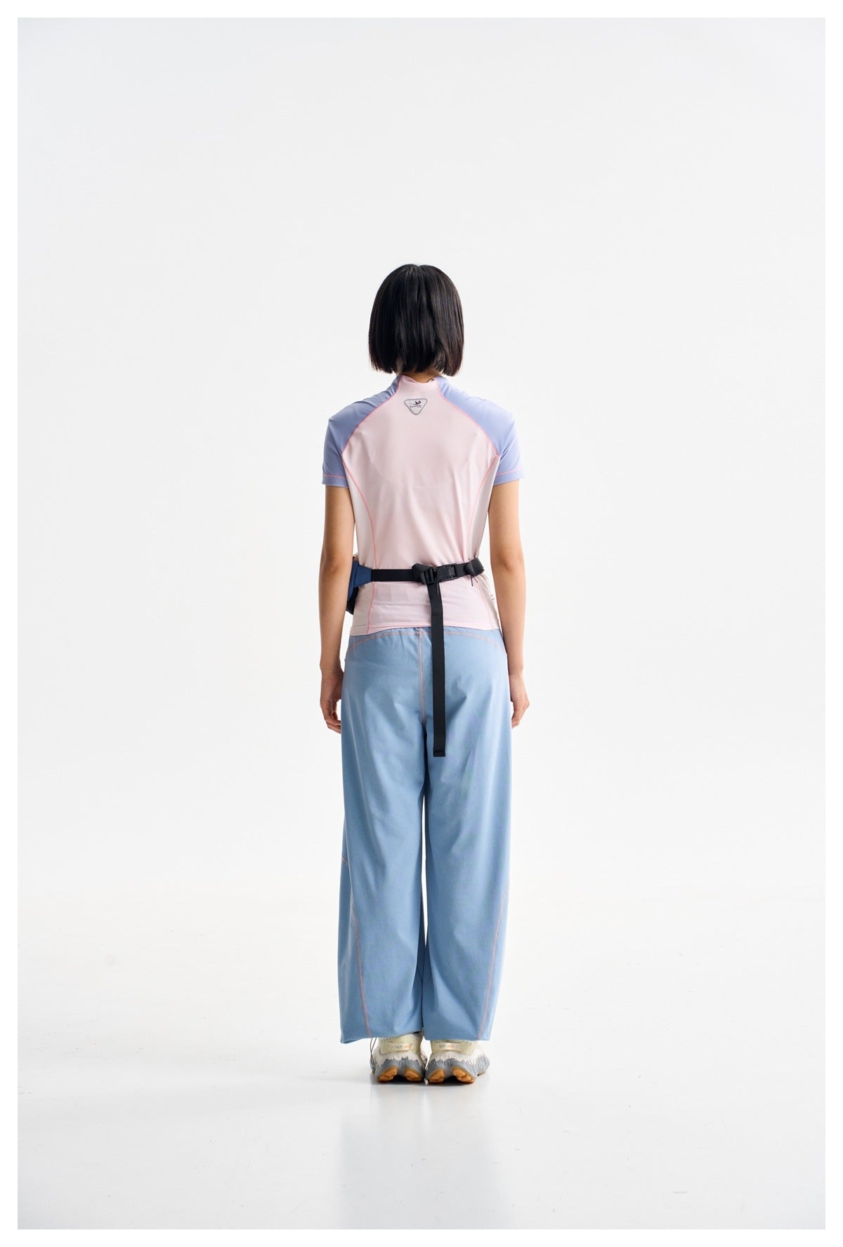 Back view of a model wearing a pale pink and blue top with loose blue trousers and a black waist belt.