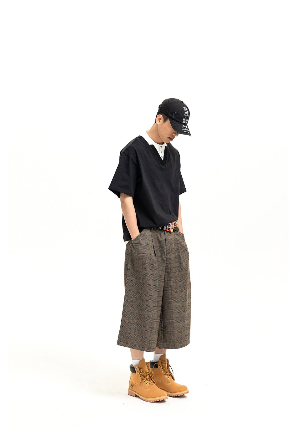 Model wearing brown-gray plaid wide-leg shorts with a black V-neck shirt, cap, and tan boots.