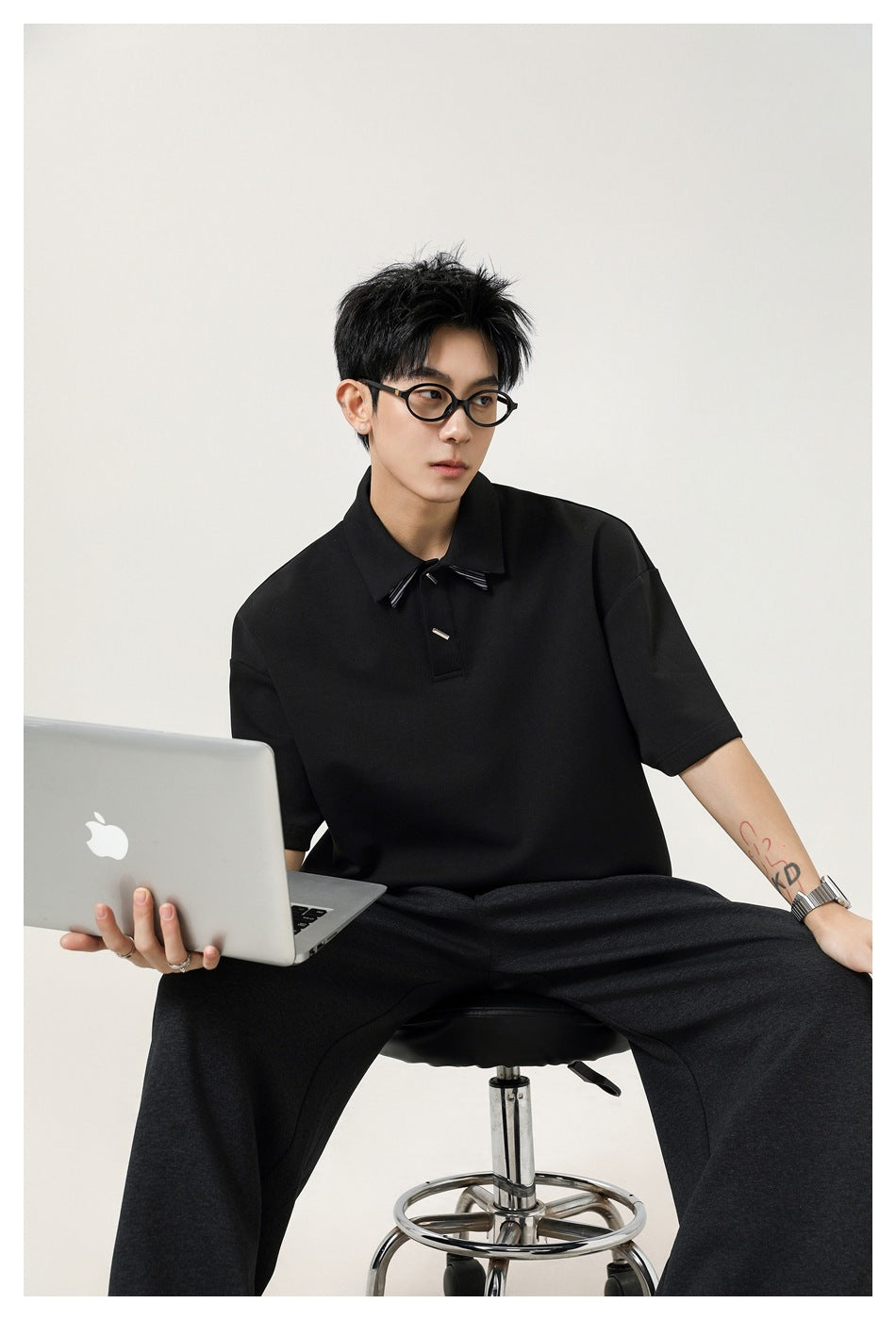 Model seated in a black oversized short-sleeve polo with pointed collar and decorative accents, holding a laptop.