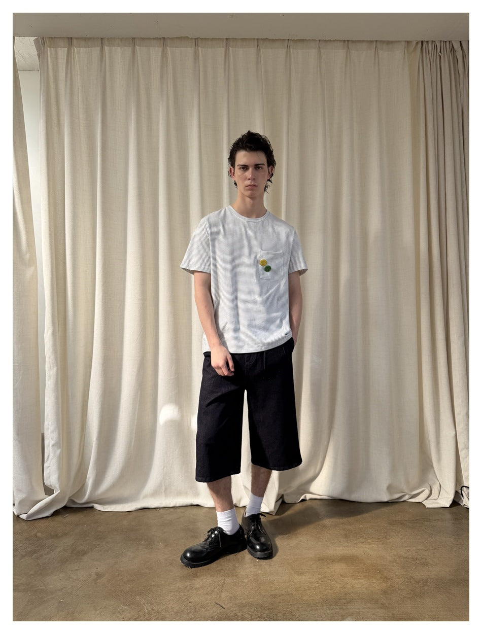 Model wearing a white T-shirt with fine gray stripes, a chest pocket and small yellow and green accents, with black shorts.