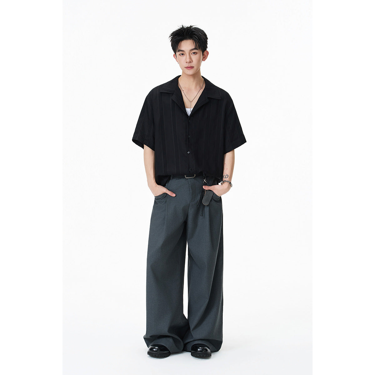 Model wearing a black short-sleeve open-collar shirt with subtle vertical stripes and charcoal wide-leg trousers.