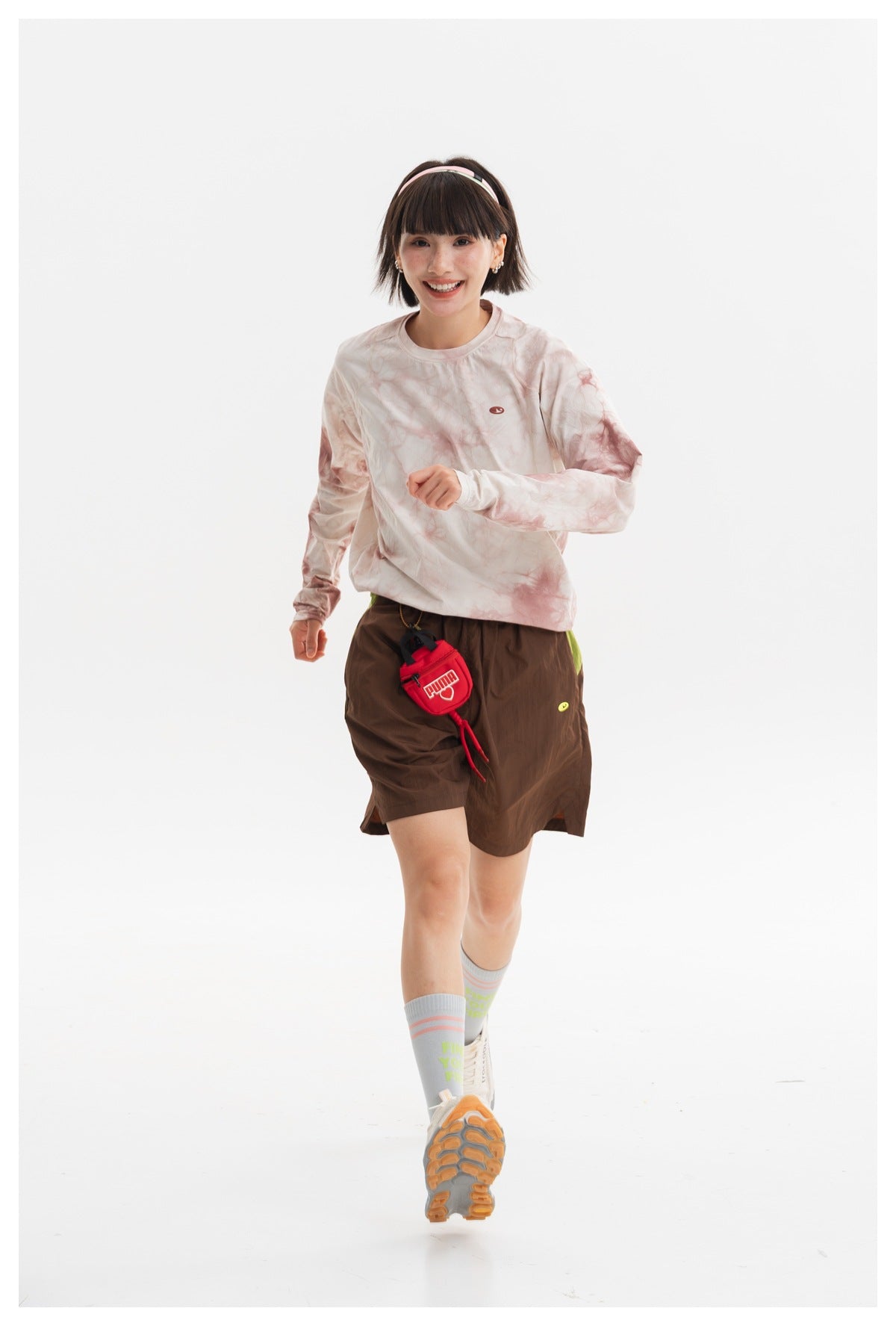 Model walking in a cream and dusty-pink tie-dye long-sleeve crewneck shirt and brown shorts, front view.