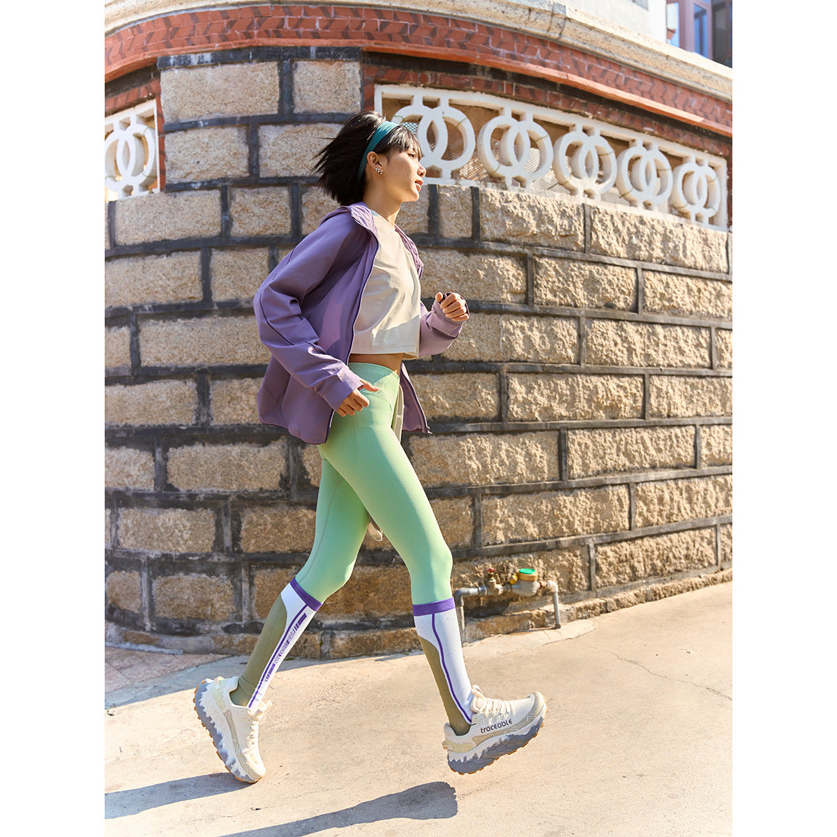 Woman jogging outdoors in a purple jacket, white cropped top, mint leggings, and white sneakers.